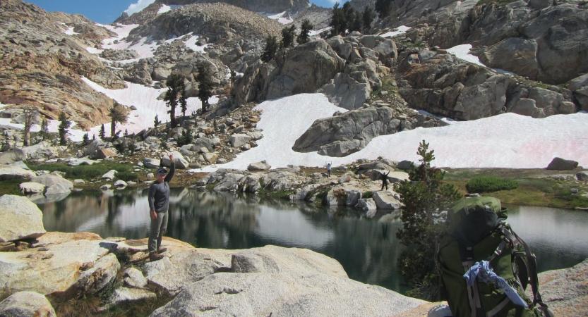 A backpack rests on a rock in the foreground, as a person stands some distance away near an alpine lake framed by boulders and snow. 